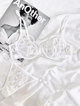 A white Jolie Unlined Transparent Lace Bra and Thong set by Petite Cherry is laid out on white fabric next to an issue of AnOther magazine, which features a black-and-white photograph of a woman on its cover. The set showcases exquisite lace detailing in a delicate circular pattern.